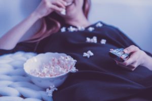 girl spending time during quarantine by laughing and eating popcorn while holding a tv remote